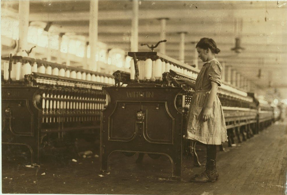 A young girl with her dark hair in a bun wearing a dress, stockings, and leather shoes monitors a bank of cotton spinning machines in the mill.